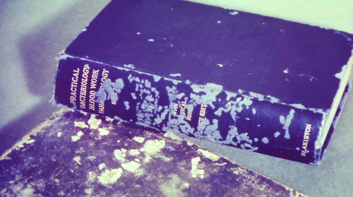 Dark cloth-bound textbook lying on another book, its spine and cover worn away in pale, irregular patches where cockroaches fed