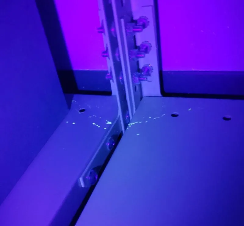Inside of a cabinet lit by a UV flashlight, with small blue-white spots and streaks glowing on the bottom shelf where it meets a slotted metal shelf upright